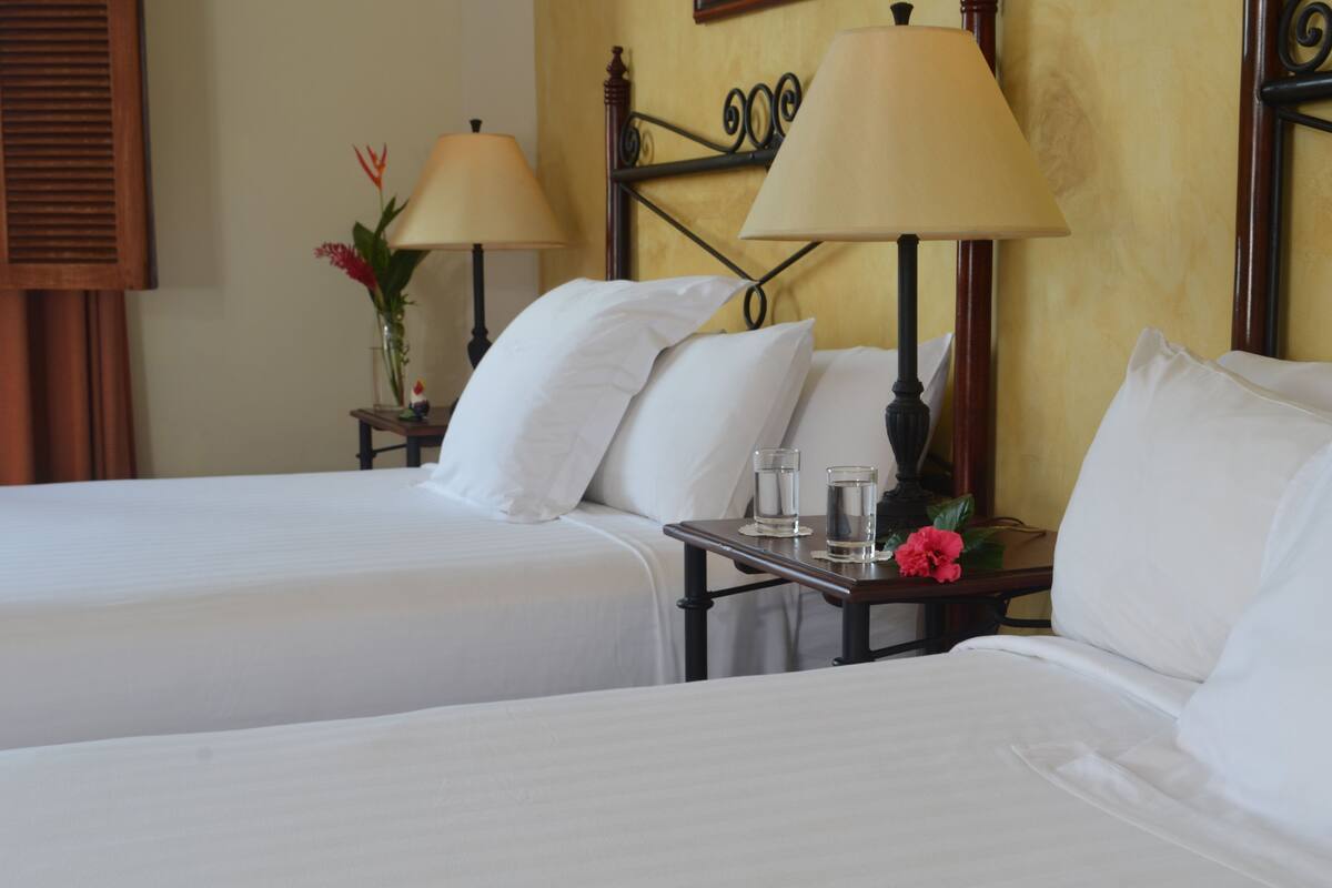 A hotel room with two beds, a nightstand with lamps, a vase with flowers, and a wall-mounted clock.