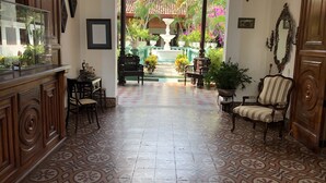 Un vestíbulo amplio con piso de baldosas estampadas, una mesa de recepción y vista a un patio con fuente y palmeras.
