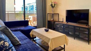Ein modernes Wohnzimmer mit einer Couch, einem Tisch mit Blumenvase, einem Fernseher und einem Balkon mit Blick auf Gebäude.