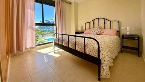 Ein Schlafzimmer mit Bett, Nachttisch, Fenster mit Vorhängen und Blick auf einen Pool sowie Gebäude.