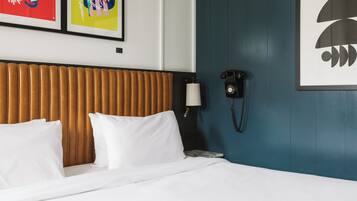 A hotel room with a bed, white pillows, a wooden headboard, a wall-mounted phone, and two framed artworks.