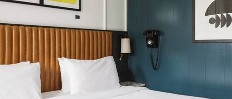 Habitación de hotel con cama, almohadillas blancas, cabecero de madera, teléfono de pared y dos obras de arte enmarcadas.