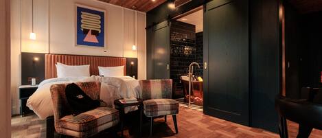 Una habitación de hotel moderna con techo de madera, una cama grande con ropa blanca, dos sillones a cuadros, una mesita pequeña y un armario de color oscuro.