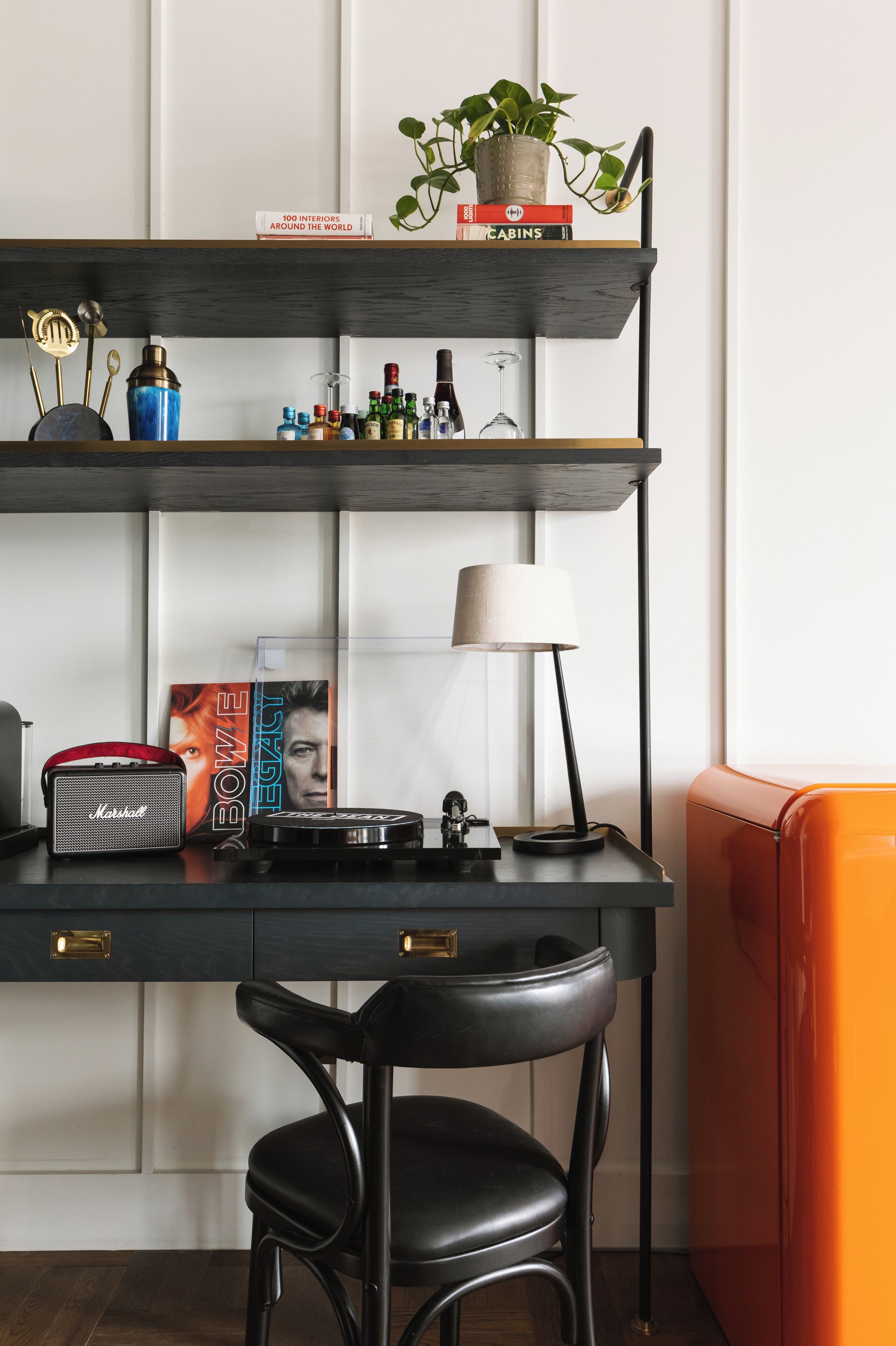A modern desk with a black chair, a lamp, a book titled 'Bowie Legacy', and a plant on a shelf.