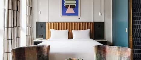 A modern hotel room with a large bed, two upholstered chairs, a small table with a plant, and a framed artwork on the wall.