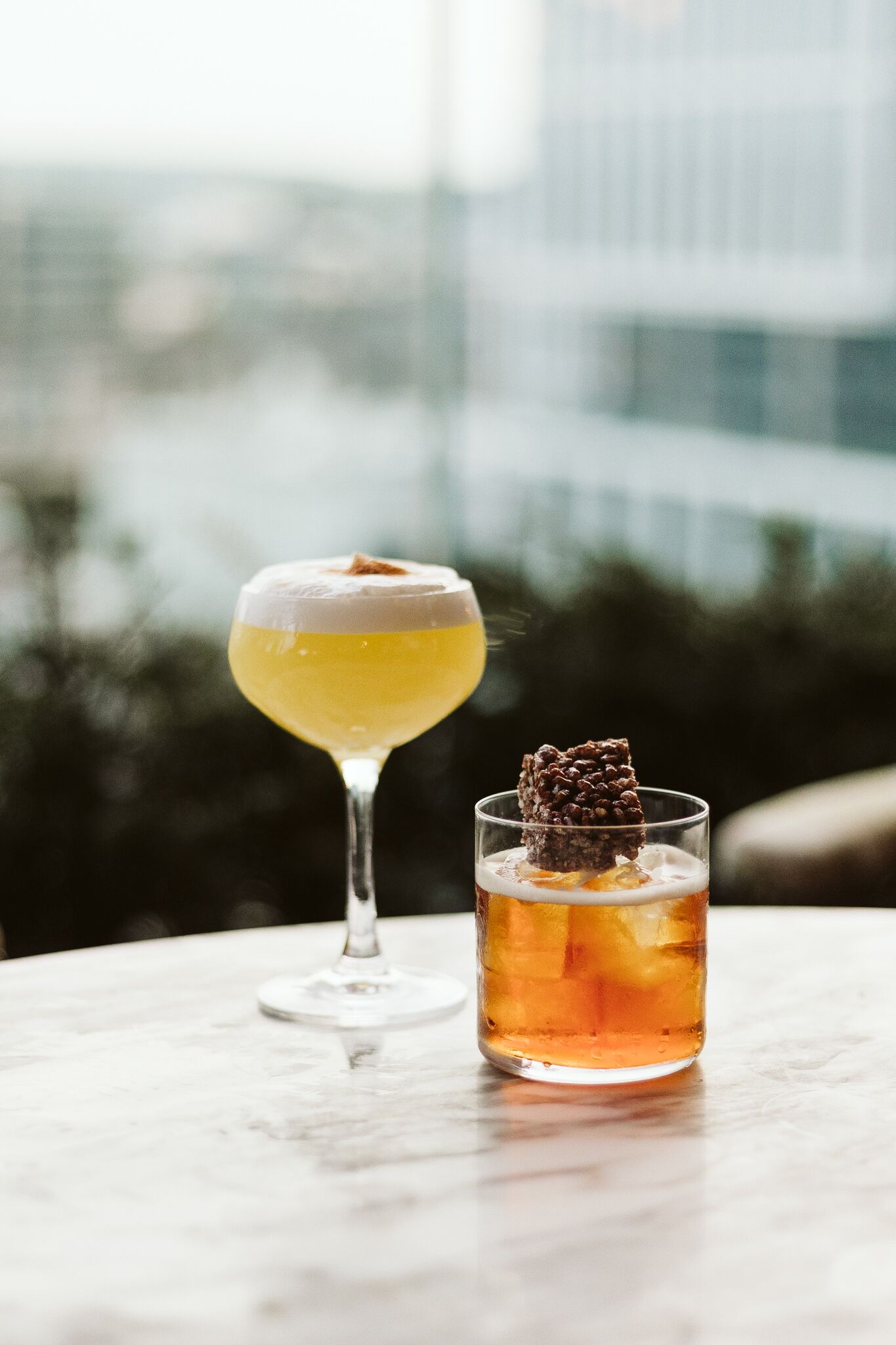 Two drinks on a table with a cityscape in the background.