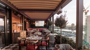 An outdoor dining area with checkered flooring, red cushioned chairs, and tables set for dining. A view of a river and cityscape is visible through large windows. - The Dean Cork (Cork)