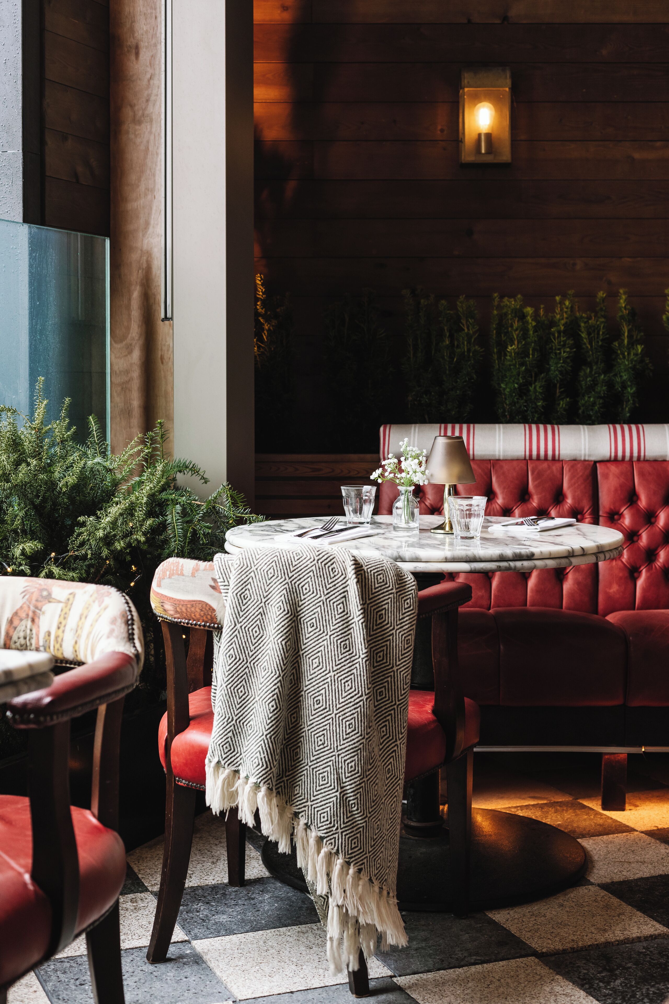 A cozy dining area with a round table set for two, a red upholstered bench, and a patterned tablecloth.