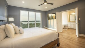 A bedroom with a large bed, a ceiling fan, and a view of a body of water through large windows. - The Oasis at Canyon Lake - Gorgeous & Modern Waterfront Home + Pool + Sweeping Lake Views (Canyon Lake)