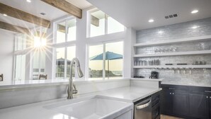 A modern kitchen with a large window, white countertops, dark cabinets, and a backsplash with a stone-like texture. - The Oasis at Canyon Lake - Gorgeous & Modern Waterfront Home + Pool + Sweeping Lake Views (Canyon Lake)