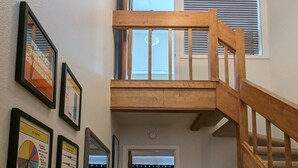 Eine Treppe führt zu einem Schlafzimmer mit Erker, das einen Ausblick nach draußen bietet. An der Wand befinden sich Bilder in Rahmen, und ein kleiner Tisch trägt eine Pflanze.