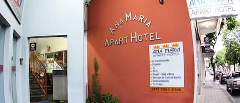 The exterior of Ana Maria Apart Hotel with a red facade, a visible entrance, and a sign listing amenities such as air conditioning, fridge, TV, Wi-Fi, and a sofa and table.