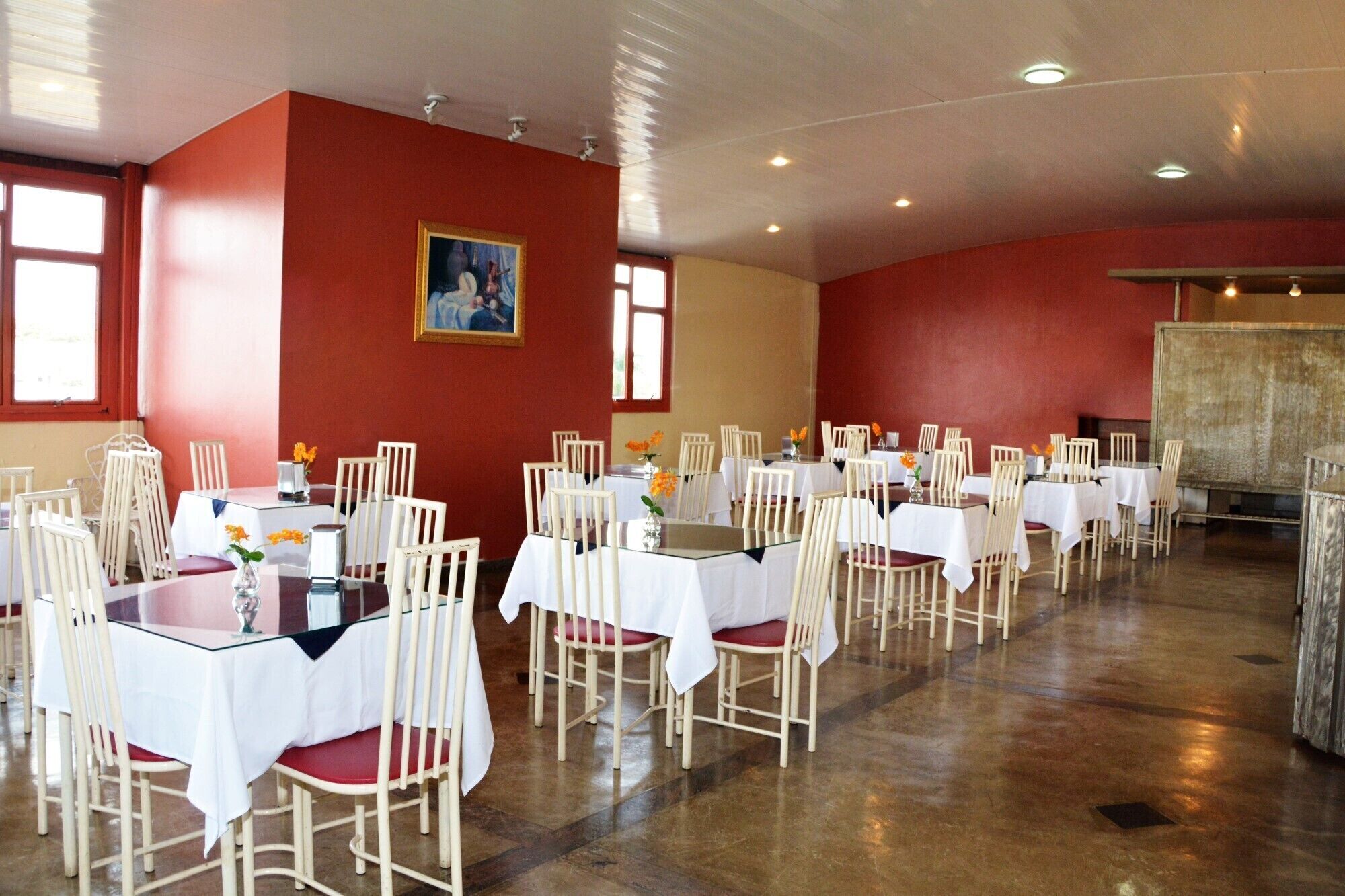 Un espace repas avec des tables et des chaises, un tableau accroché au mur et une grande fenêtre.