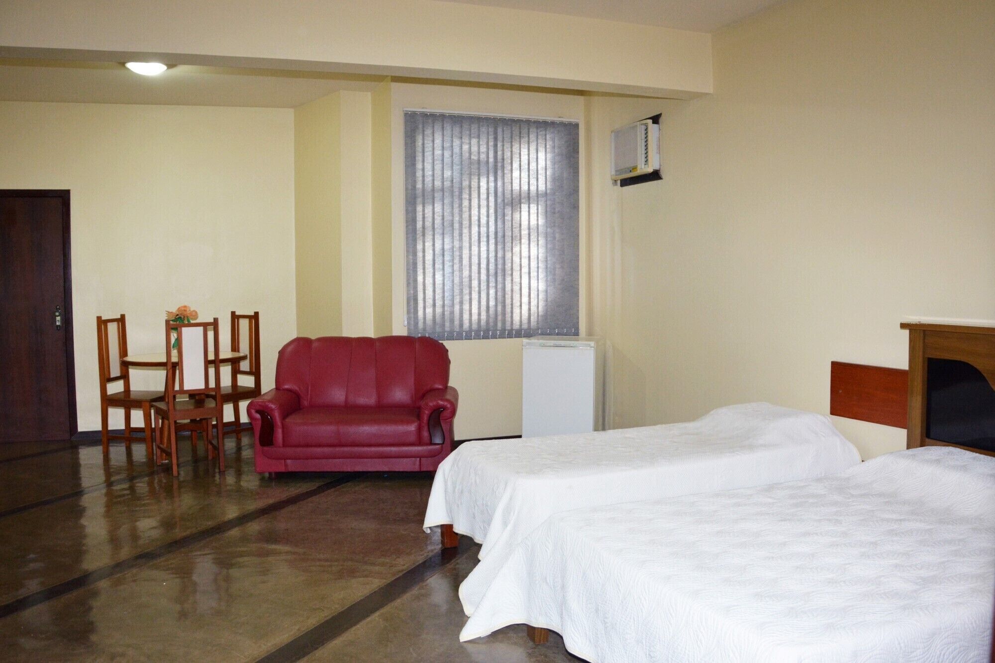 Une chambre d’hôtel avec deux lits simples, un fauteuil rouge, une petite table avec un vase et une fenêtre avec des stores.