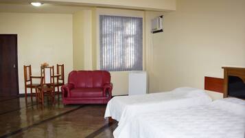 Une chambre d’hôtel avec deux lits simples, un fauteuil rouge, une petite table avec un vase et une fenêtre avec des stores.