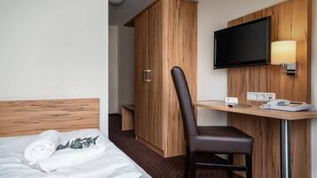 A hotel room with a bed, a wooden wardrobe, a desk with a chair, a television, and a lamp.