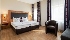 A hotel room with a double bed, a nightstand with a lamp, a chair, and a sofa. - ClassicX Landhaus & Hotel (Gensingen)