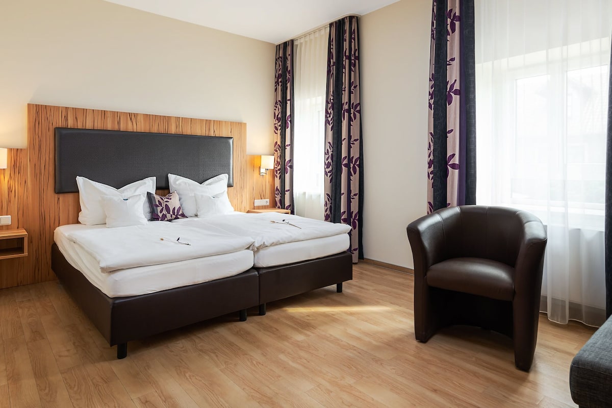 A hotel room with a double bed, bedside lamps, a chair, a sofa, and a window with curtains.