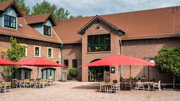 Un bâtiment en briques surmonté d’un toit en tuiles, avec une terrasse dotée de parasols rouges et une grande fenêtre ornée de volets verts.