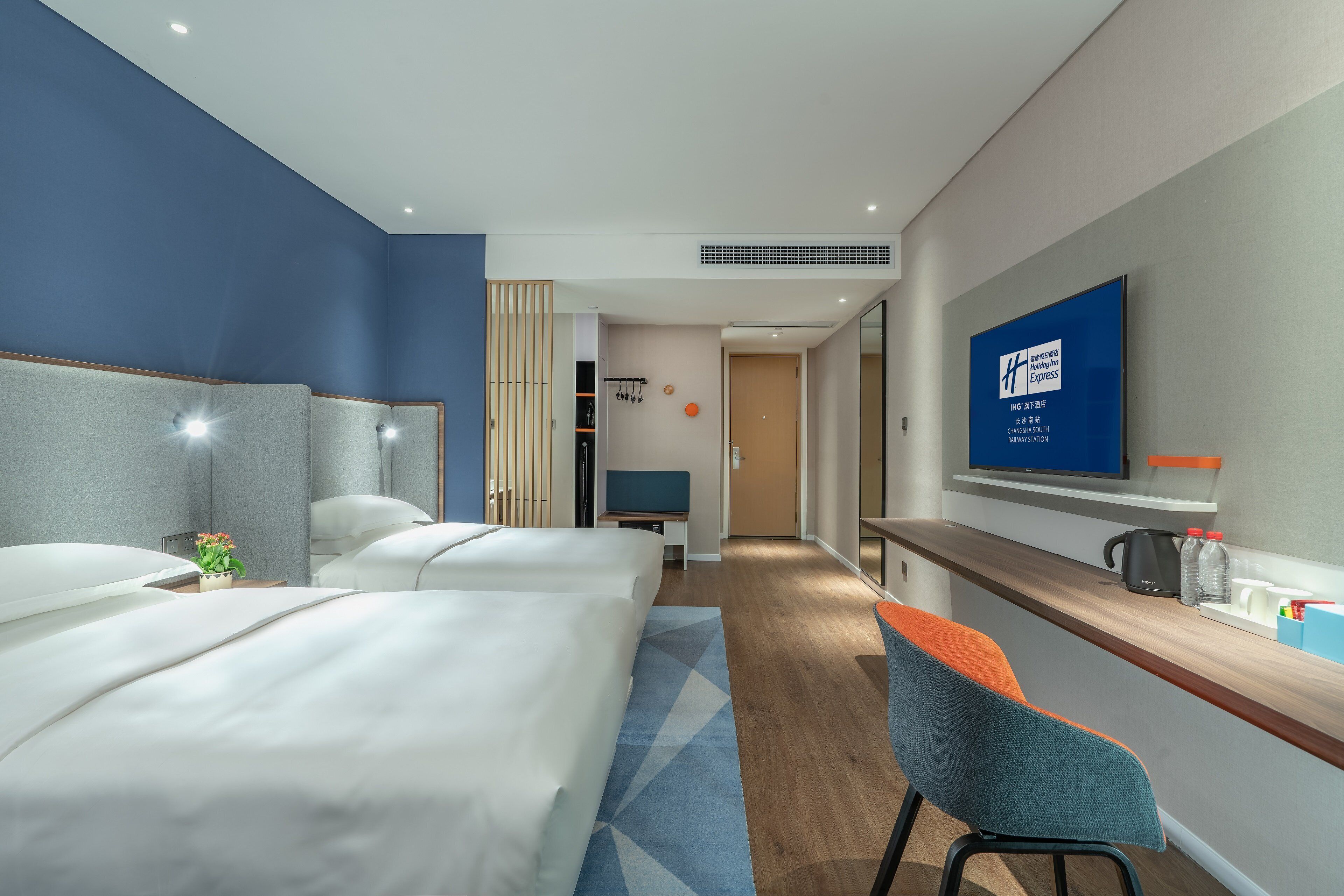 Habitación de hotel con dos camas, un escritorio, una silla, televisión y vista al pasillo.