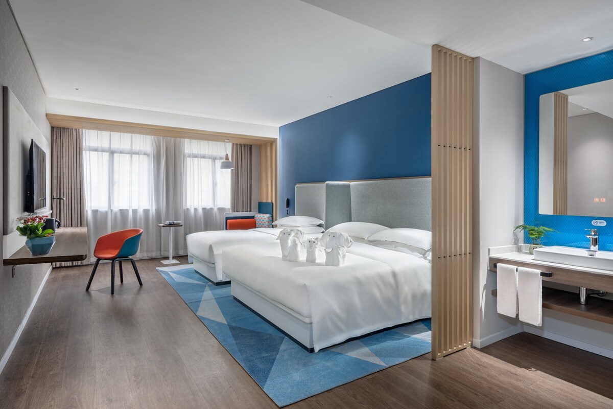 Una habitación de hotel moderna con una cama grande, un escritorio, una silla, un televisor y un baño con espejo y lavamanos.