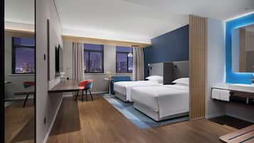 Habitación de hotel con cama, escritorio, silla, mesita, mesita de noche, espejo y vista de la ciudad por la noche.
