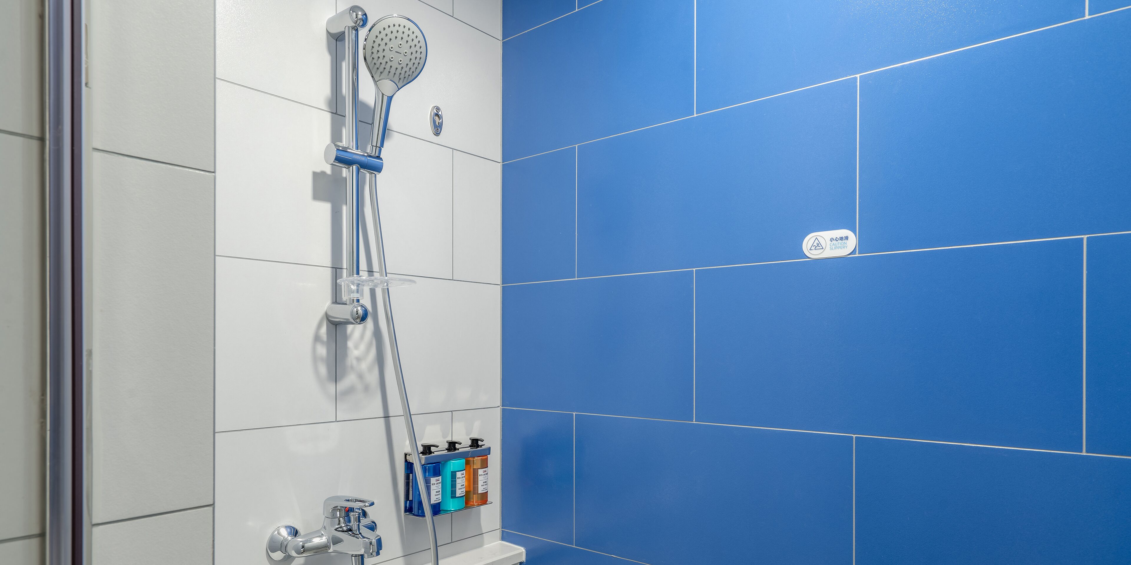 A shower with a handheld showerhead and a wall-mounted control. There are two bottles of shampoo and conditioner on a shelf below the shower.
