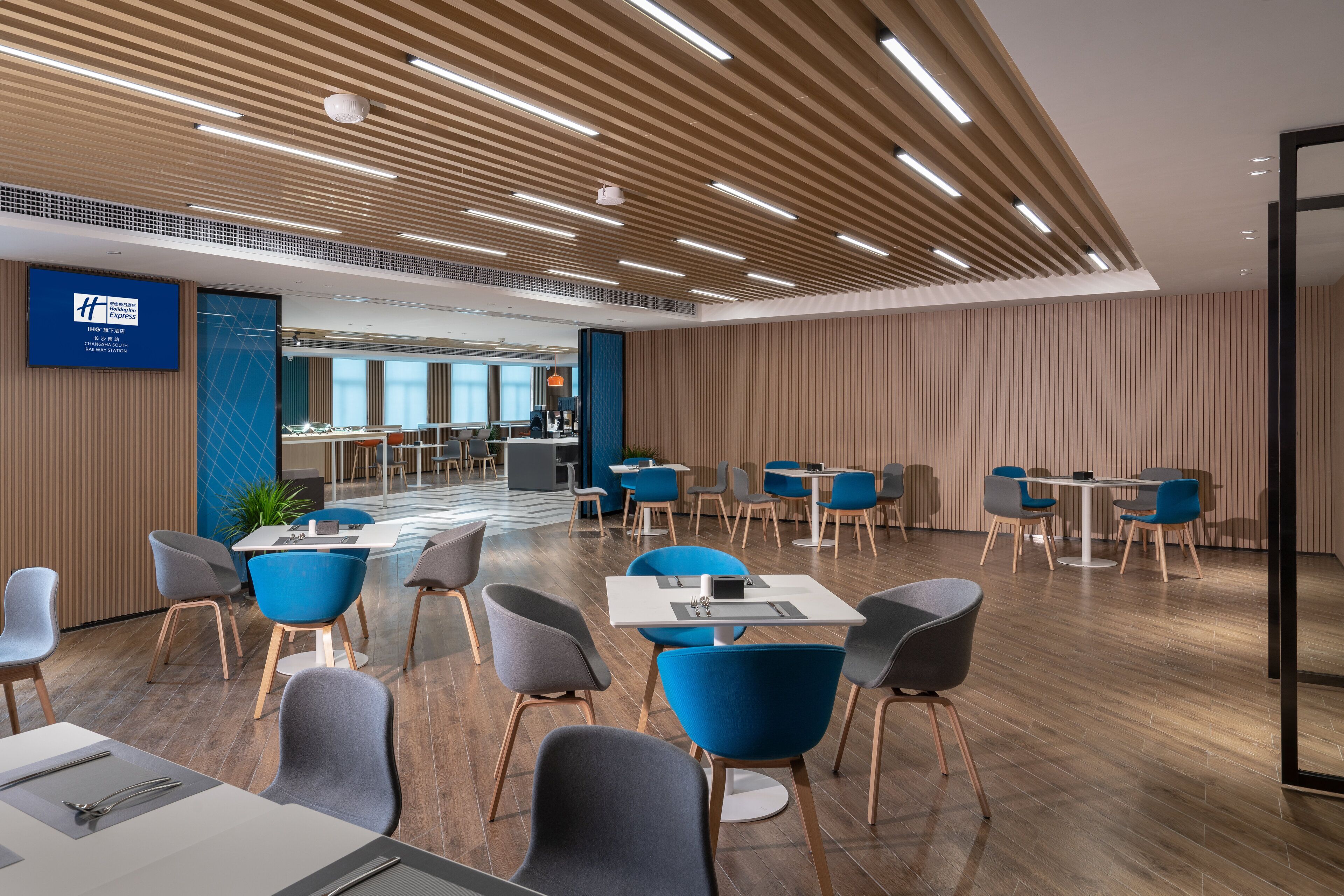 A modern hotel lobby with a high ceiling, wooden paneling, and a mix of blue and grey seating areas.