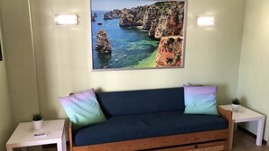 Une pièce comprenant un canapé en bois, deux petites tables, un tableau représentant un paysage côtier accroché au mur et une plante en pot posée sur l’une des tables.