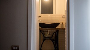 A narrow hallway leading to a bathroom with a sink, mirror, and a hanging light fixture. - Toledo Class (Naples)