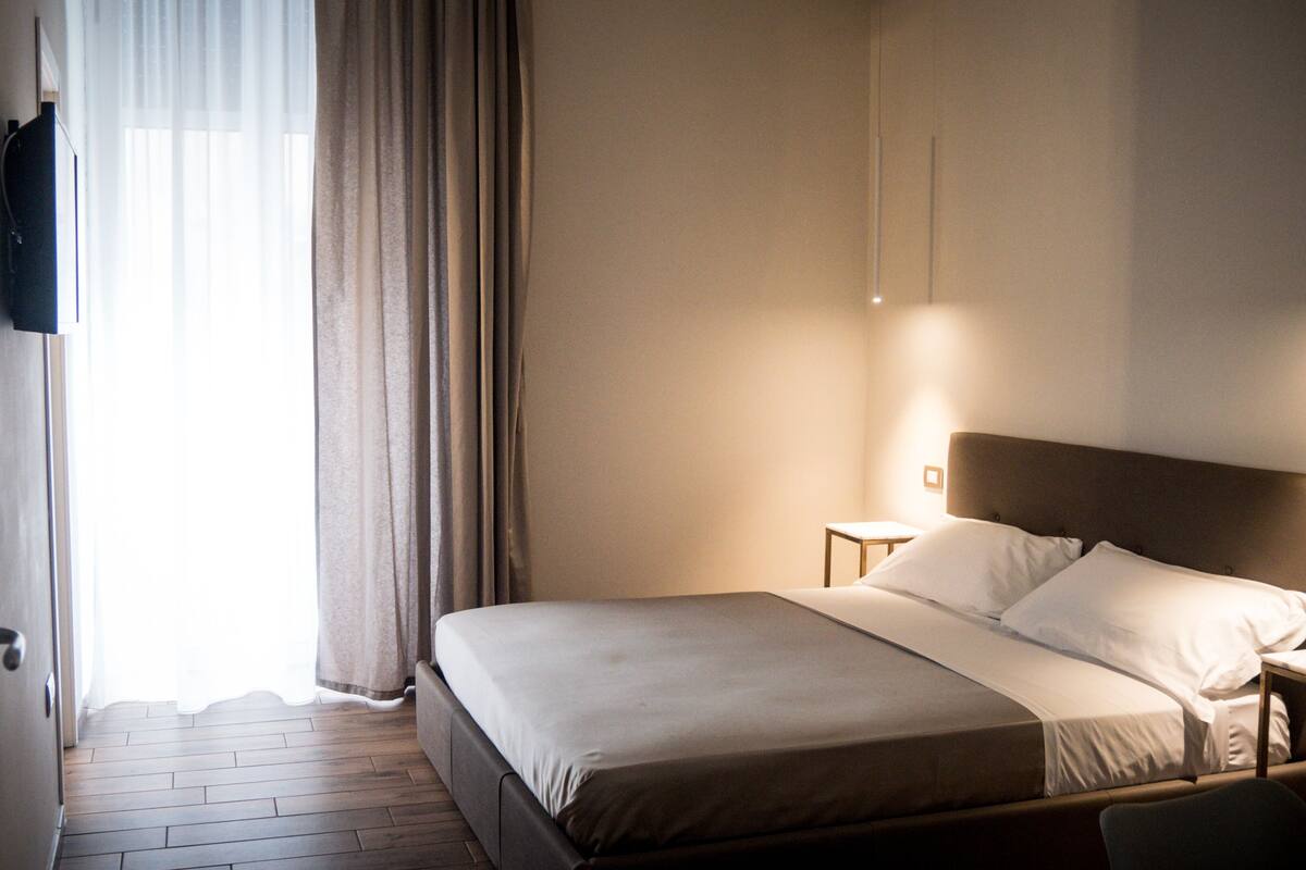 Habitación de hotel con cama, mesita de noche, televisor montado en la pared y ventana con cortinas.