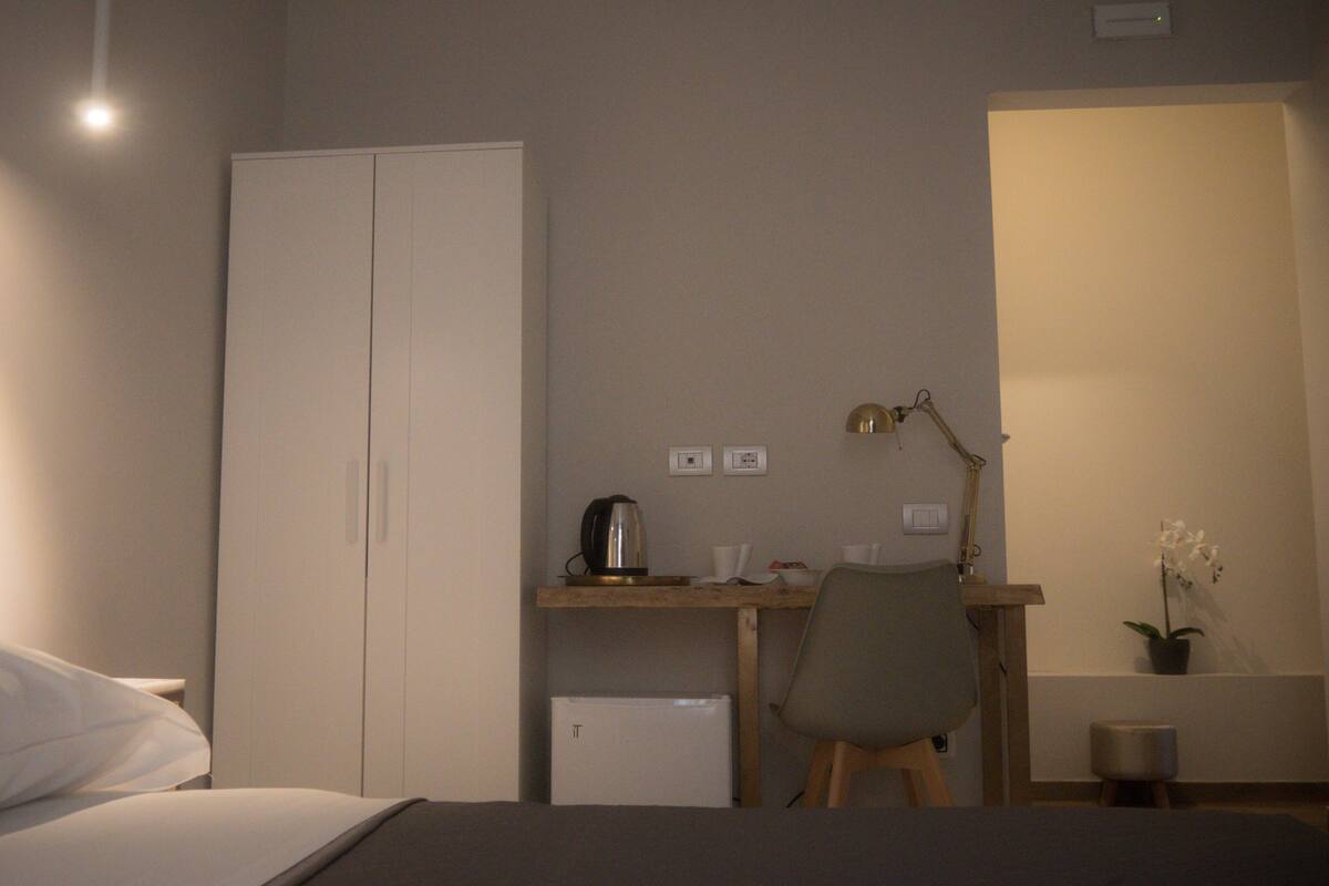 Un dormitorio con una cama, un escritorio con una silla, un armario, una lámpara de escritorio y una pequeña planta en maceta.