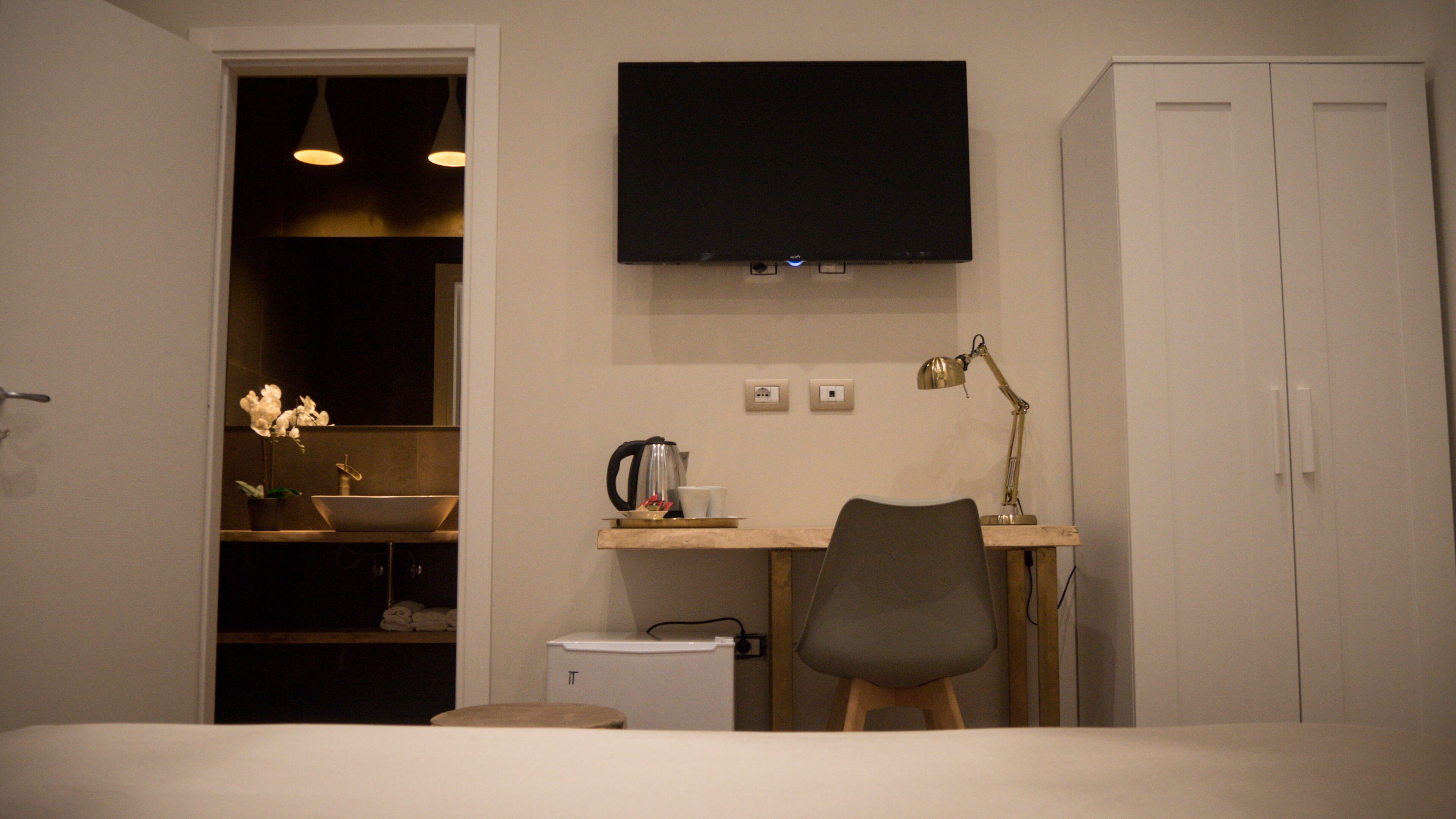 A small, modern room with a bed, a desk with a chair, a TV, a wardrobe, and a bathroom with a sink and a mirror.