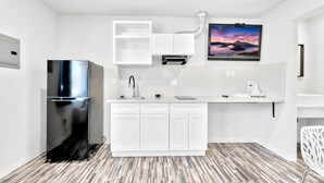 A compact kitchen with white cabinetry, a stainless steel refrigerator, a built-in microwave, and a flat-screen TV mounted above the counter. - Tradewinds Motel (Lakewood)
