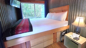 A wooden-framed bed with white linens, a red and black patterned rug, a television mounted on the wall, and a bedside table with a lamp and a thank you card.
