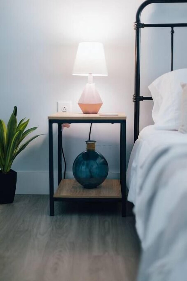 Uma cama com roupa de cama branca, uma mesa de cabeceira com abajur, uma planta em vaso e um vaso sobre a mesa.