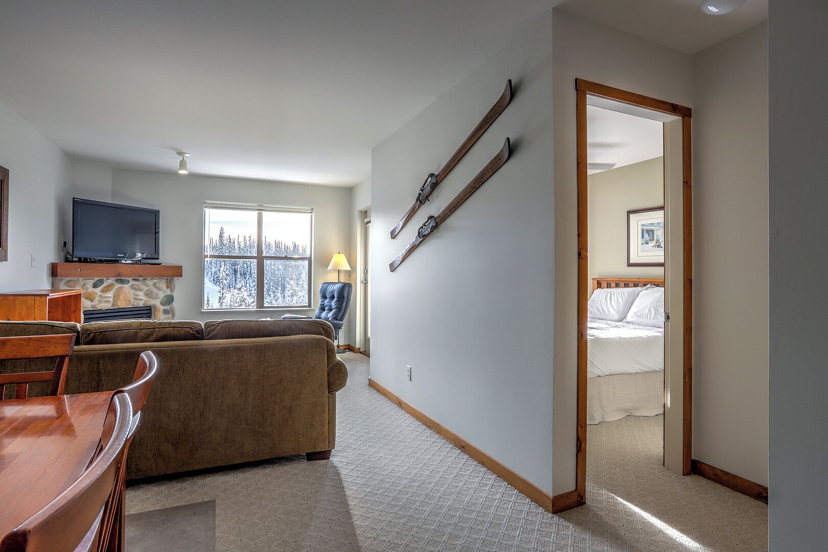 A cozy living room with a sofa, TV, and fireplace, a dining area with a table and chairs, and a bedroom with a bed and a door leading to a snowy view.