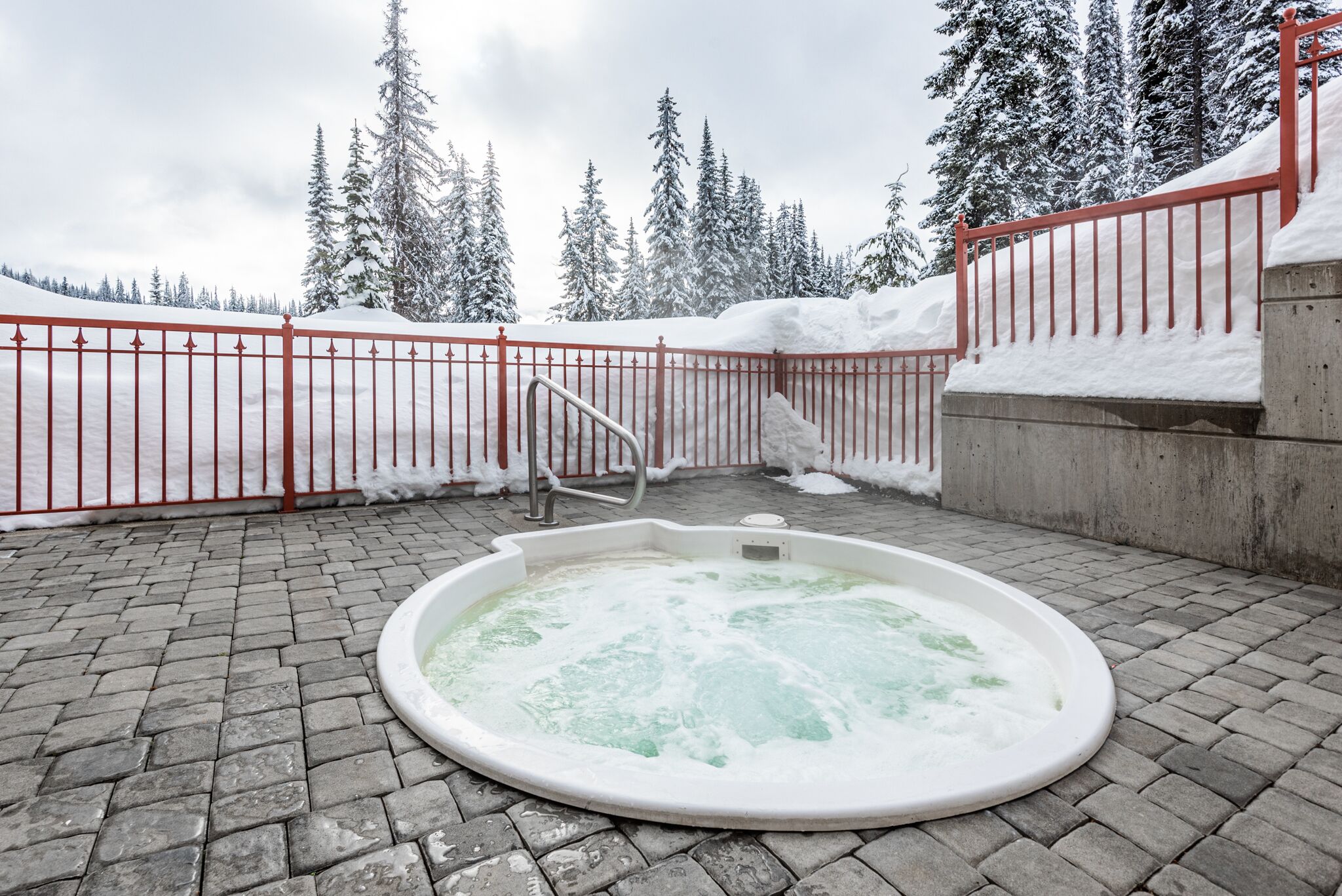 A hot tub in a snowy outdoor area with a red fence and snow-covered trees in the background.
