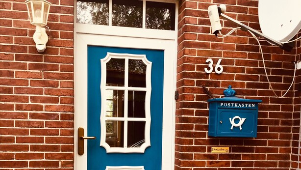 Une porte bleue avec des bordures blanches, une boîte aux lettres portant l’inscription « POSTKASTEN », et le numéro « 36 » sur le mur.
