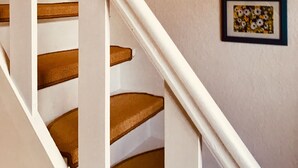 Un escalier avec des marches en bois et des rampes blanches, menant à un palier où sont accrochées des œuvres d’art encadrées au mur.