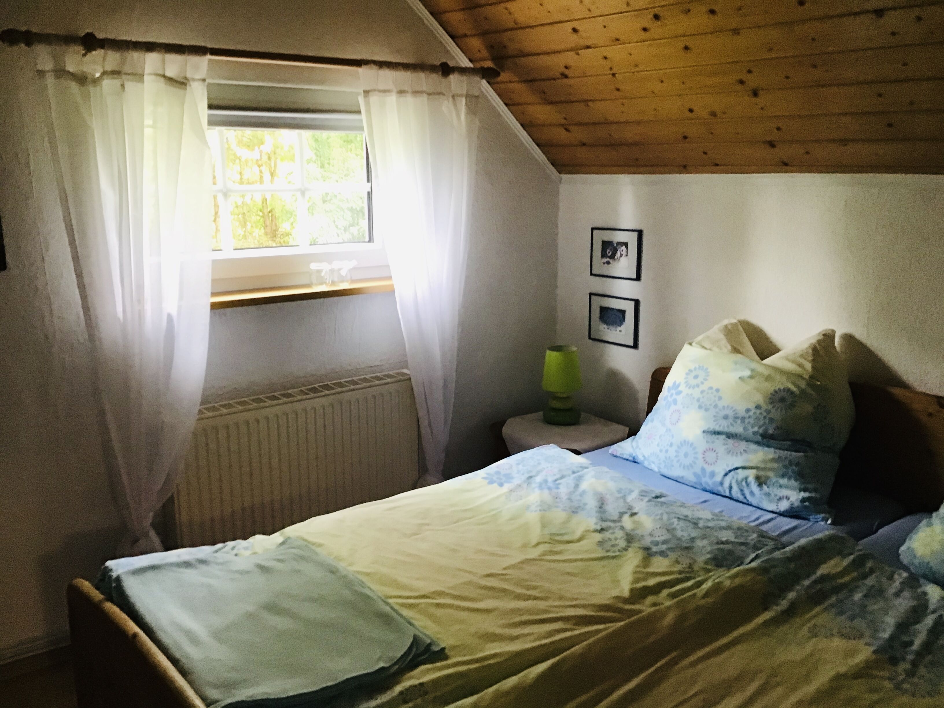 A bedroom with a bed, a window with curtains, a radiator, a bedside table with a lamp, and two framed pictures on the wall.