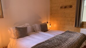A neatly made bed with a quilted blanket and pillows, a bedside table with a lamp, and a wooden wall with a window. - A gorgeous 3 bed apartment, 3 minutes walk from the centre of Samoëns (Samoëns)