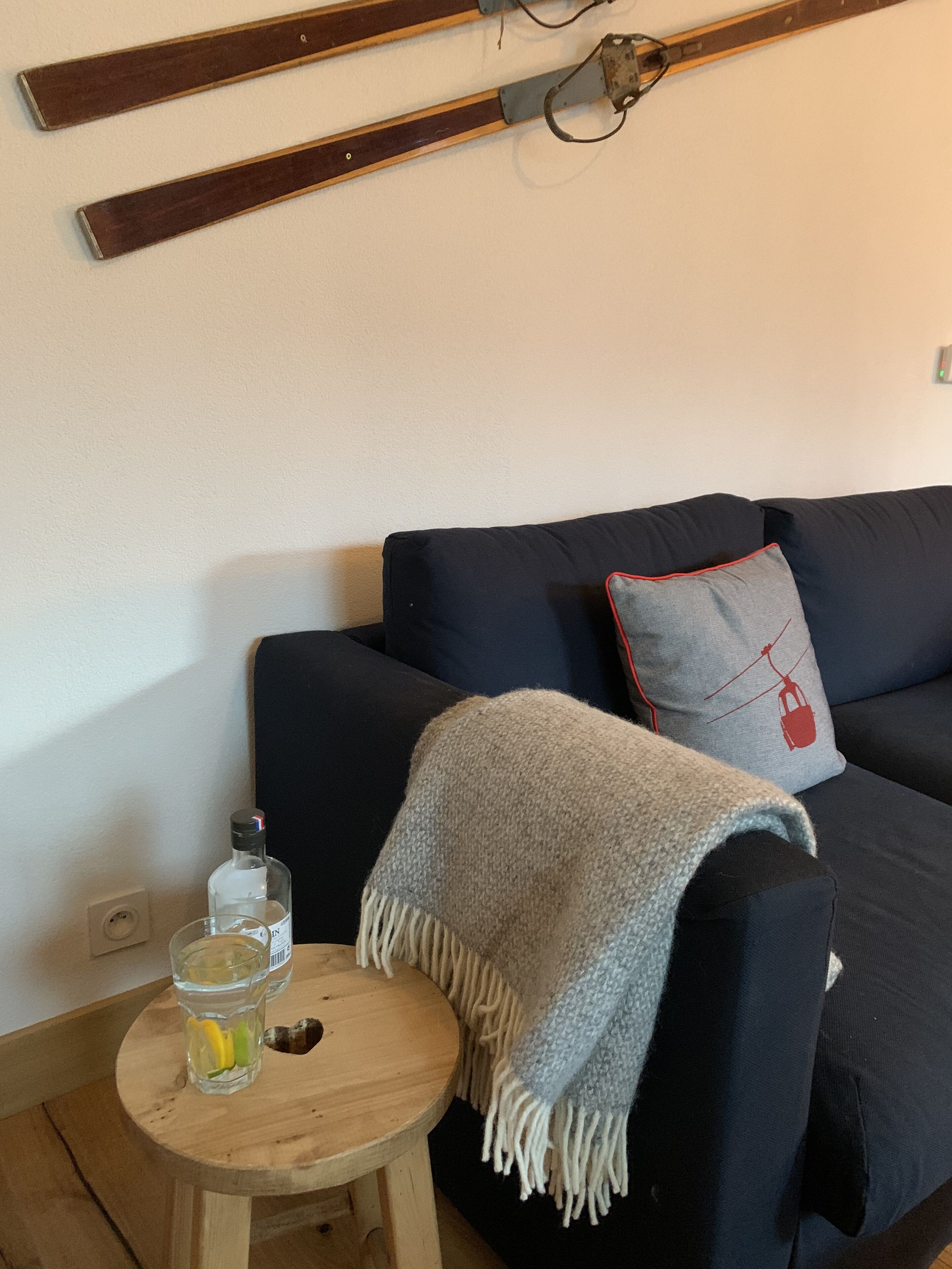 A living room with a dark blue sofa, a small round wooden table with a glass of water and a bottle, and a grey blanket draped over the sofa.