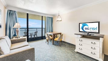 Quarto de hotel com varanda, televisão, mesa de jantar com cadeiras e cômoda branca.