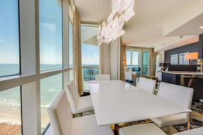 A hotel room with a large white table, white chairs, a view of the ocean, and a modern chandelier.