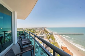 A balcony with wicker chairs, a glass railing, and a view of a beach and pier.