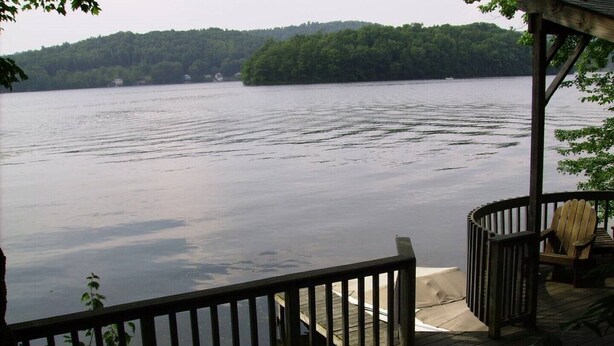 A wooden deck with a railing overlooking a calm lake, surrounded by trees. - Expansive Lakehouse with Jacuzzi (Argyle)