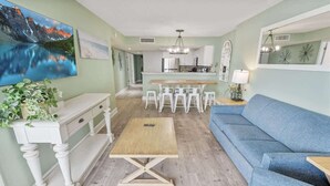 A living room with a blue sofa, a wooden coffee table, and a dining area with white chairs. - Sand Dunes Resort North Tower, 17th Floor, Pools! (Myrtle Beach)