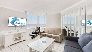 A modern living room with a white TV, a sofa, a coffee table, and a mirrored wall. - Oceanfront Corner, Low Floor, Ocean Annies, Pools! (Myrtle Beach)