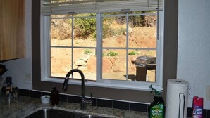 Une cuisine avec une fenêtre donnant sur un jardin, un évier et un plan de travail sur lequel se trouvent divers objets.